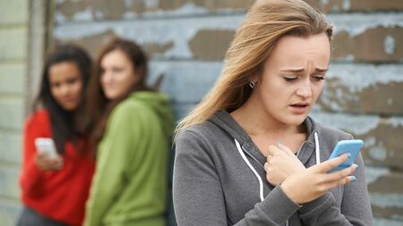 Teenage girl staring at phone with a hurt look as 2 girls talk behind her back