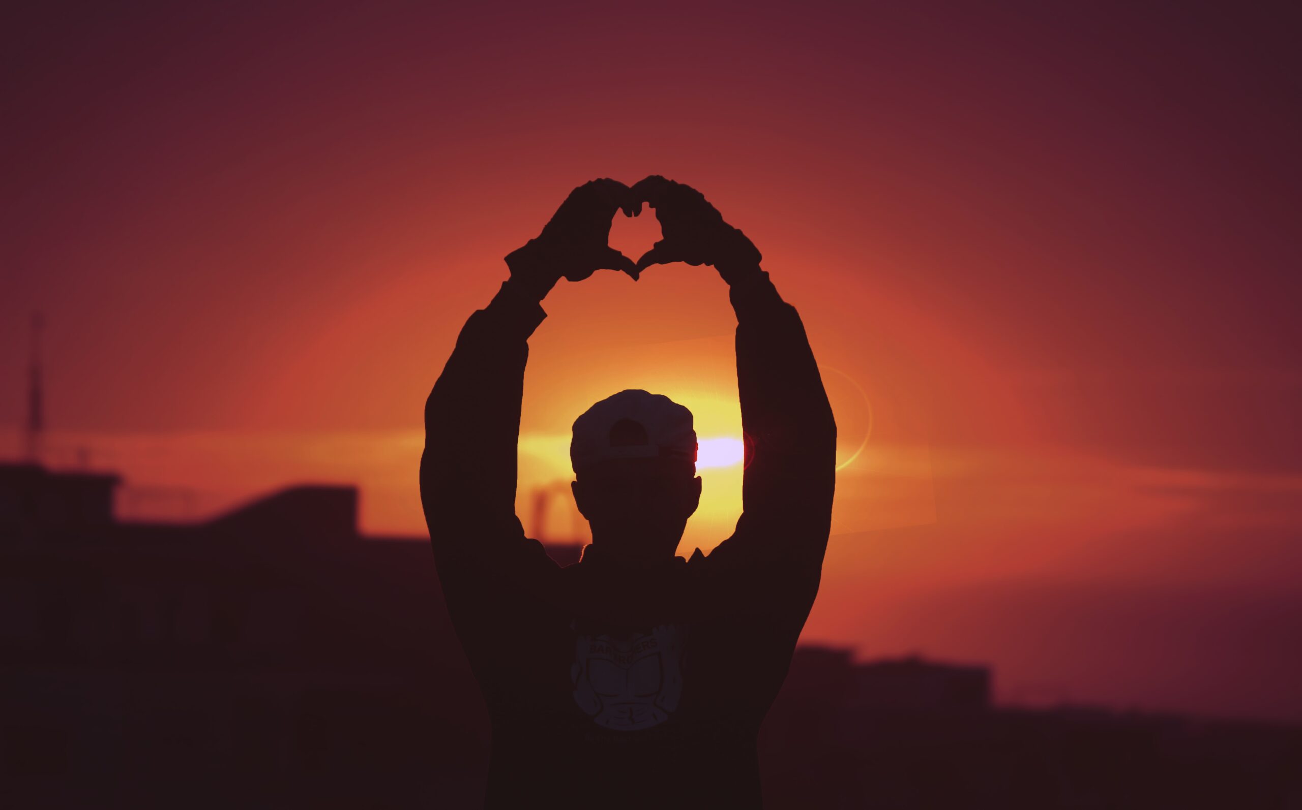 man holding up a heart with his hands in the sunset